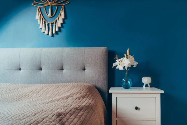 Cozy bedroom with dark blue walls and Boho home interior decor: macrame wall hanging decoration, white bedside table,vase with lilly flower, aroma lamp. Dark modern stylish room. Selective focus