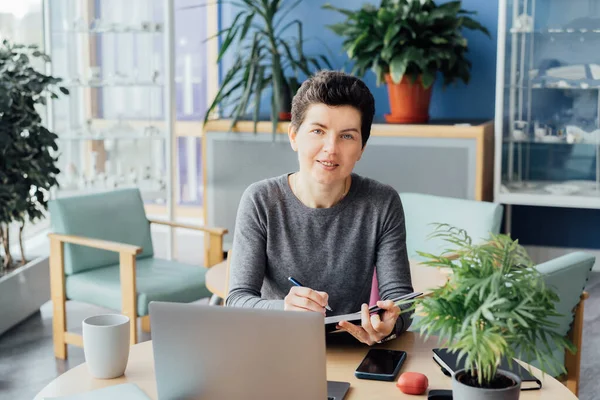 Portrait of smiling middle aged neutral gender business woman interviewing person online, using laptop, making notes in her paper notebook. Online video call, IT HR specialist, recruiting process
