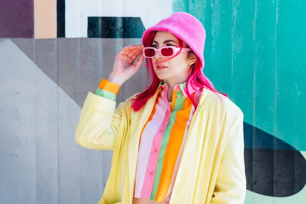 Urban spring street fashion look. Vanilla Girl. Kawaii vibes. Candy colors design. Bucket hat trends. Young woman with pink hair and sunglasses in multicolor outfit on the outdoor wall background.