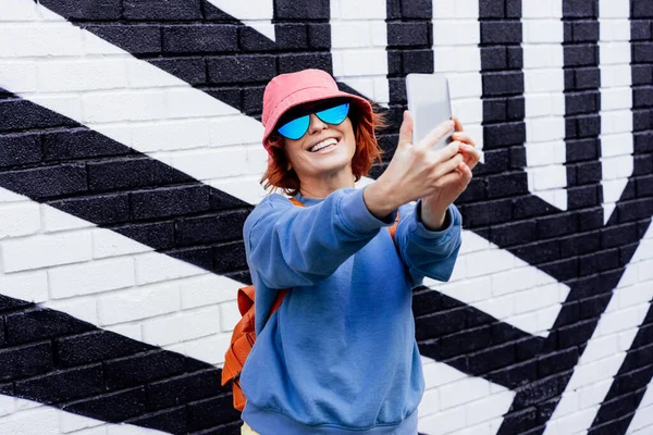 Excited redhead woman taking a selfie photo outdoors. Emotional hipster fashion woman in bright clothes, heart shaped glasses, bucket hat taking selfie photo on the phone camera. Street fashion.