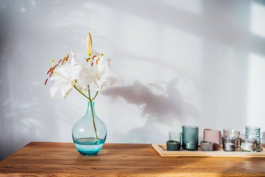 Modern minimalist İskandinav tarzı iç tasarımı beyaz poster modeli, mumlar ve beyaz zambak çiçekleri güneş ışığı altındaki ahşap bir konsolda ve gri duvarda ev bitkileri gölgeleri. Seçici odak