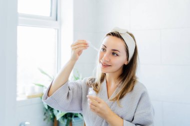 Young woman in bathrobe looking in the mirror, holding dropper with hyaluronic acid serum close to face and smiling. Skin hydrating. Cosmetic spa procedures. Beauty self-care at home. Selective focus