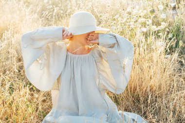 Açık renkli elbisesi ve samandan şapkası olan, elinde çimen sapı tutan ve gün batımında yüksek çayırlarda eğlenen narin, tanınmamış bir kadın. Doğada bir yaz akşamı. Sakin ve uyumlu. Tatil