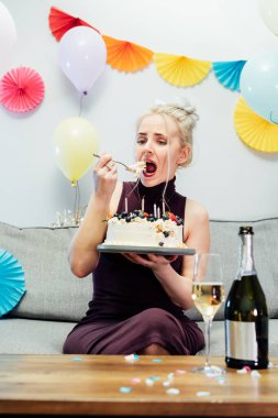 Üzgün, üzgün kadın kokteyl elbisesi içinde ve parti şapkası takmış kanepede oturup doğum gününü kutlarken pasta yiyor. Doğum gününü yalnız kutluyor, Kendi Partisi, sıkıcı parti. Seçici odak.