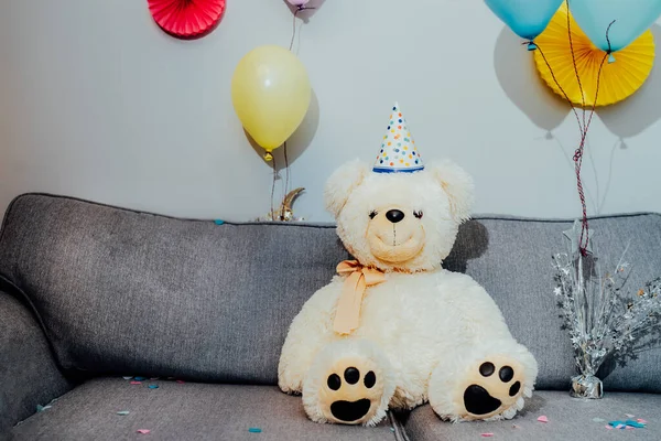 Big Teddy bear with balloons in party cap sitting on the gray sofa in decorated room. Birthday or festive holiday background. Selective focus, copy space.