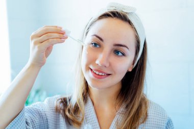 Young woman in bathrobe looking in the mirror, holding dropper with hyaluronic acid serum close to face and smiling. Skin hydrating. Cosmetic spa procedures. Beauty self-care at home. Selective focus