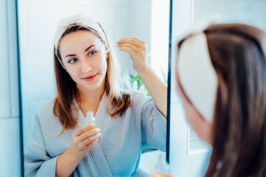 Young woman in bathrobe looking in the mirror, holding dropper with hyaluronic acid serum close to face and smiling. Skin hydrating. Cosmetic spa procedures. Beauty self-care at home. Selective focus