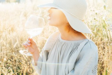 Açık renkli elbisesi ve hasır şapkası olan, elinde şarap kadehiyle, yüksek çayırlarda gün batımını seyreden, tanınmamış bir kadını yakından çek. Doğada yaz akşamının tadını çıkar. Sakin ve uyumlu. Tatilde. Seçici odak