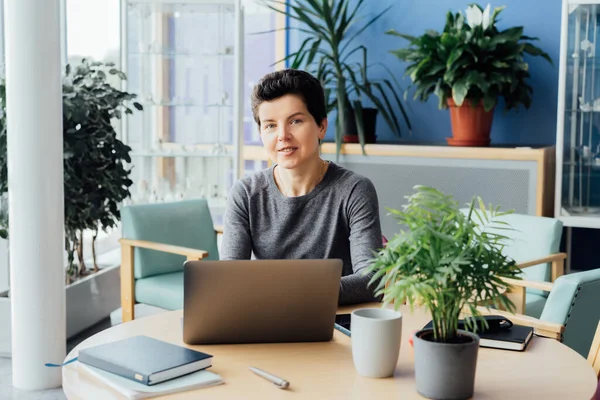 Neutral gender middle aged woman with no make-up in casual clothing using laptop and smiling while working indoors in her workstation in an open space office. Online video call, IT HR, recruiting.