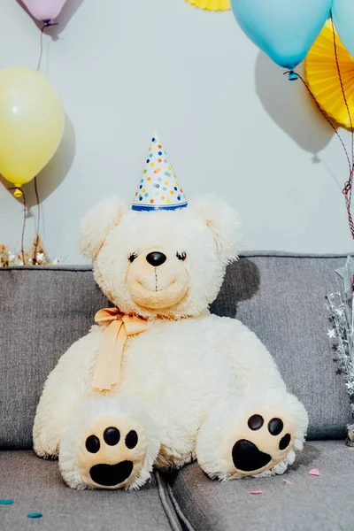 Big Teddy bear with balloon in party cap and sitting on the gray sofa in decorated room. Birthday or festive holiday vertical background. Selective focus, copy space.