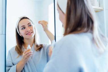 Young woman in bathrobe looking in the mirror, holding dropper with hyaluronic acid serum close to face and smiling. Skin hydrating. Cosmetic spa procedures. Beauty self-care at home. Selective focus