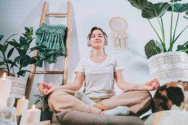Sağlıklı genç bir kadın evde meditasyon yapıyor, kulaklık takıyor, modern Scandi 'nin evindeki minderde lotus pozisyonunda oturuyor. Meditasyon, rahatlama, gözler kapalı. Seçici odak.