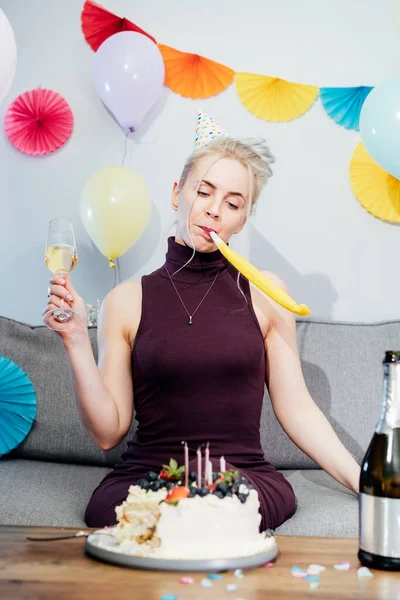 Sad, upset woman in festive dress and party cap blowing in birthday pipe and drinking champagne while sitting on the sofa with festive cake. Celebrates birthday alone. Bored party. Vertical card