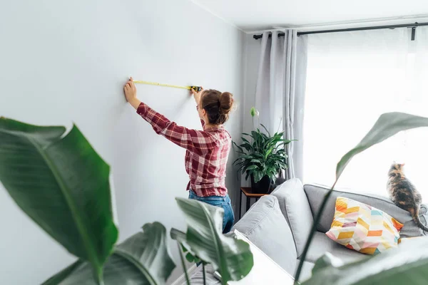 stock image Young woman doing measuring with a measure tape on the wall. Girl wants to put a picture on the wall at home. Housekeeping work. Doing repair herself. DIY, gender equality in work concept