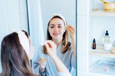 Young woman in bathrobe looking in the mirror, holding dropper with hyaluronic acid serum close to face and smiling. Skin hydrating. Cosmetic spa procedures. Beauty self-care at home. Selective focus