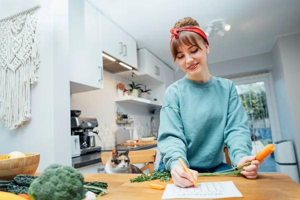 Young Woman Filling Veganuary Calendar While Eating Fresh Carrot Sitting — Stock Photo, Image