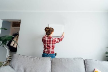 Arkaya bakan genç bir kadın duvara resim, resim çerçevesi asıyor. Yeni şık dairesinde rahat bir hayat süren sıradan bir kadın. Genç bayan dekorasyon odası. Evi taşıma ve yenileme