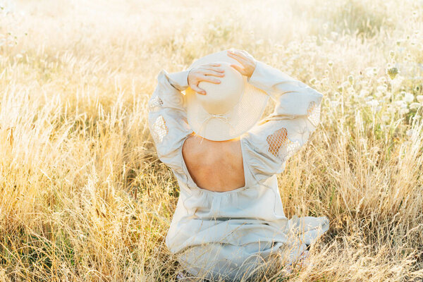 Tender back view woman in light airy dress with open back and straw hat enjoying the moment in a high grass meadow at sunset. summer evening in nature. Calm, harmony. Freckles on body, natural beauty