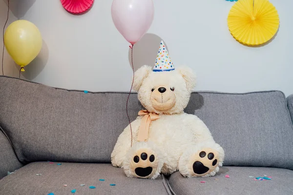 Big Teddy bear with balloons in party cap sitting on the gray sofa in decorated room. Birthday or festive holiday background. Selective focus, copy space.