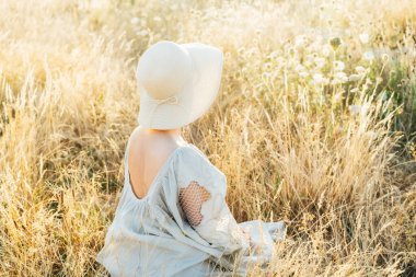 Açık sırtı ve hasır şapkası olan, açık hava görünümlü yumuşak bir kadın günbatımında yüksek çim çayırında anın tadını çıkarıyor. Doğada bir yaz akşamı. Sakin, ahenkli. Vücudundaki çiller, doğal güzellik.