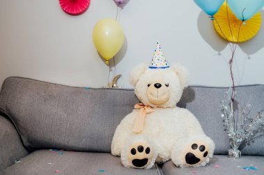 Parti şapkalı büyük oyuncak ayı dekore odasındaki gri koltukta oturuyor. Doğum günü ya da bayram geçmişi. Seçici odak, boşluğu kopyala.