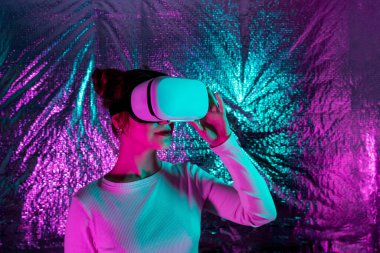 Meta evren konsepti. Beyaz elbiseli ve VR gözlüklü gülümseyen kadın renkli neon ışığı olan folyo desenli bir arka planda kulaklıkla sanal ve artırılmış gerçekliği araştırıyor. Seçici odak