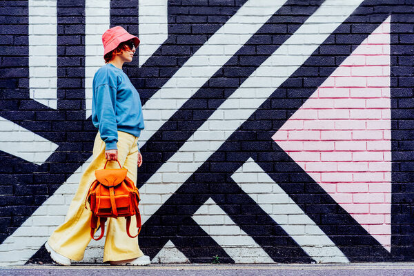Hipster fashion young woman in bright clothes, sun glasses, bucket hat with backpack walking on the background of the painted brick wall. Urban city street fashion. Selective focus. Copy space