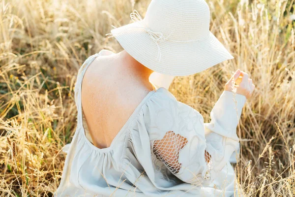 柔嫩的后视镜女人穿着轻飘飘的衣服 开著背 戴着草帽 日落时分在高高的草地上享受着这一刻 在大自然的夏夜 身体上的雀斑 自然美 — 图库照片