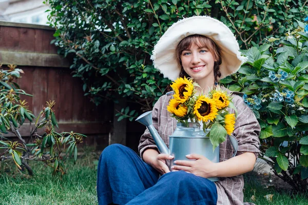 stock image Portrait of smiling young female farmer woman holding watering can with fresh sunflowes bouquet while sitting in the green garden background. Cottagecore lifestyle. Selective focus, copy space