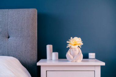 Kapalı beyaz komodinin yanında seramik kadın şeklinde vazo, gül çiçeği ve koyu mavi duvar zemininde mumlar. Modern tarz kadın yatak odası. Evde kendini iyileştirme yeri. Seçici odak.