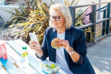 Orta yaşlı bir iş kadını salatalı hamburger yiyor ve parkta piknik masasında oturup telefon kullanıyor. Kilo vermek için sağlıklı beslenme çantası. Dengeli diyet