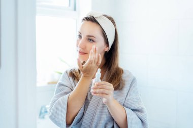 Bornozlu genç bir kadın aynaya bakıyor, elinde hyaluronik asit serumuyla şişeyi tutuyor ve yüzüne uyguluyor. Cilt sulanır. Kozmetik spa prosedürleri. Evde kendine çok iyi bakıyorsun. Seçici odak.