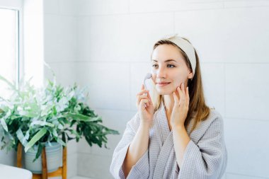 Bornozlu genç bir kadın aynaya bakıyor ve banyoda yeşim taşıyla yüz masajı yapıyor. Cilt bakımı için doğal prosedürler kavramı. Ev Güzeli kendi kendine bakım tedavisi. Seçici odak