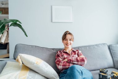 Mutlu bir şekilde gülümseyen genç bir kadın evindeki duvara bir resim astıktan sonra koltukta dinleniyor. Oda temizliği işi. Kendini tamir ediyor. DIY, cinsiyet eşitliği kavramı. Ev dekorasyonu.