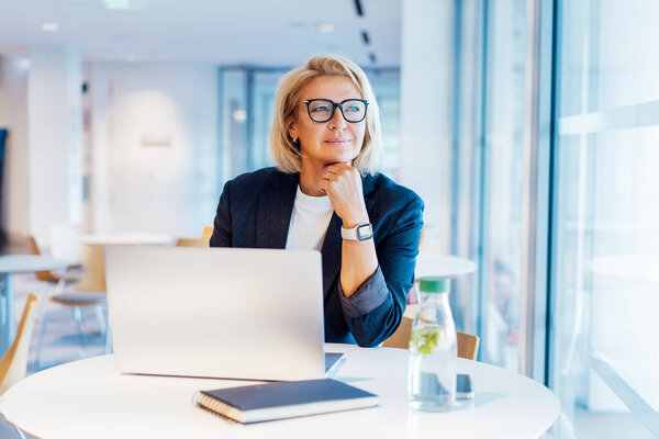 50s confident mature businesswoman dreamy looking at window, middle-aged experienced senior female professional working on laptop in open space office. Female entrepreneur working remotely