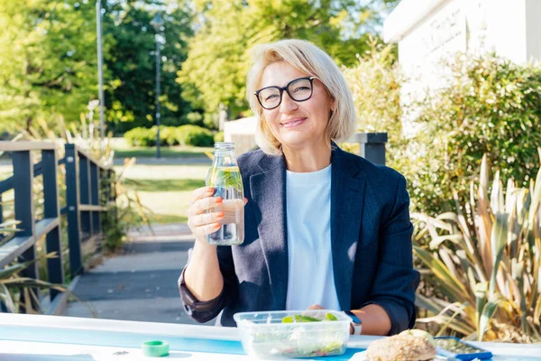 Healthy habit to drink water. Smiling middle aged business woman with bottle of water with lemon and mint during her outdoor lunch break. Control body hydration, Tracker water balance. Healthy living