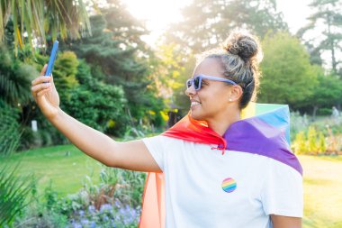 Gökkuşağı bayrağına sarılmış, LGBT topluluğunun sembolü olan genç Latin mutlu kadın, bir şehir parkındaki bir gurur kutlamasında cep telefonuyla selfie çekiyor. İnsan hakları, eşitlik, LGBT, Özgürlük ve Mutluluk