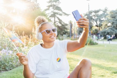 Gökkuşağı bayrağına sarılmış, LGBT topluluğunun sembolü olan genç Latin mutlu kadın, bir şehir parkındaki bir gurur kutlamasında cep telefonuyla selfie çekiyor. İnsan hakları, eşitlik, LGBT, Özgürlük ve Mutluluk.