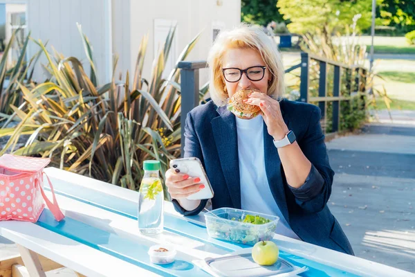 stock image Middle-aged business woman eating a wholemeal burger with salad and using a phone sitting at picnic table in the park while having a break. Healthy lunch box for weight loss. Balanced diet.
