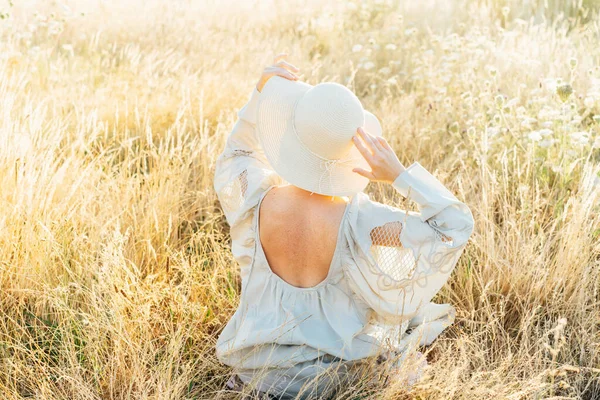 Tender back view woman in light airy dress with open back and straw hat enjoying the moment in a high grass meadow at sunset. summer evening in nature. Calm, harmony. Freckles on body, natural beauty.