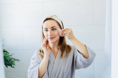 Bornozlu genç bir kadın aynaya bakıyor ve tuvalette gua sha roller ile yüz masajı yapıyor. Cilt bakımı için doğal prosedürler kavramı. Ev Güzeli kendi kendine bakım tedavisi. Seçici odak