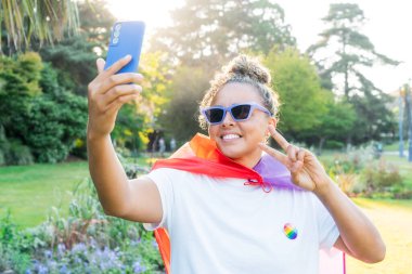 Gökkuşağı bayrağına sarılmış, LGBT topluluğunun sembolü olan genç Latin mutlu kadın, bir şehir parkındaki bir gurur kutlamasında cep telefonuyla selfie çekiyor. İnsan hakları, eşitlik, LGBT, Özgürlük ve Mutluluk.