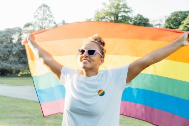 Açık havada gökkuşağı bayrağı sallayan melez bir kadın. Genç lezbiyen aktivist gülümsüyor ve Lgbtq sosyal hareketinin bayrak sembolünü taşıyor. Eşitlik, özgürlük ve insan hakları için savaş. Onur Ayı Kutlaması.