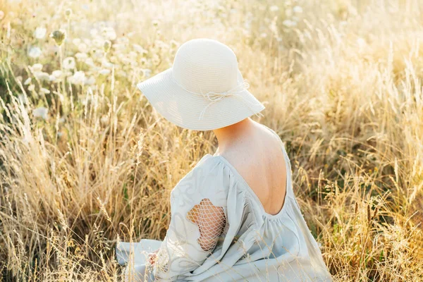 夕暮れ時に芝生の高い牧草地で瞬間を楽しむオープンバックとわら帽子と軽い風通しの良いドレスの公開バックビューの女性 自然の中で夏の夜 落ち着いて調和を 体のそばかす 自然の美しさ — ストック写真