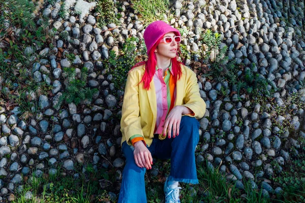 Urban spring street fashion look. Vanilla Girl. Kawaii vibes. Candy colors design. Bucket hat trend. Young woman with pink hair and sunglasses in multicolor outfit on the stones and plants background.