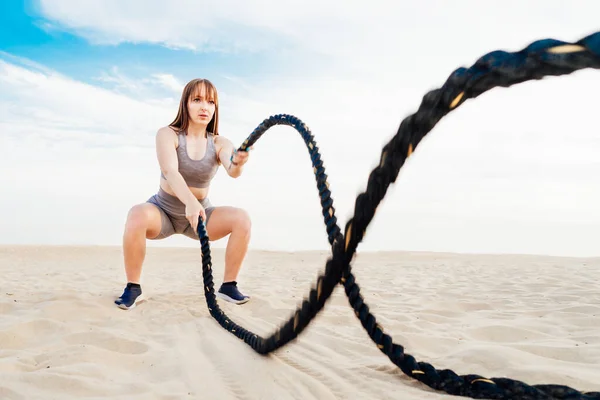 Muscular young concentrated woman working out with battle ropes on the beach, intense functional circuit training. Doing sport outdoors. Crossfit, fitness and workout concept. Selective focus