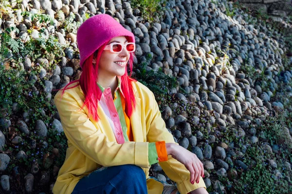 Urban spring street fashion look. Vanilla Girl. Kawaii vibes. Candy colors design. Bucket hat trend. Young woman with pink hair and sunglasses in multicolor outfit on the stones and plants background.