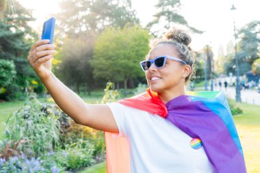 Gökkuşağı bayrağına sarılmış, LGBT topluluğunun sembolü olan genç Latin mutlu kadın, bir şehir parkındaki bir gurur kutlamasında cep telefonuyla selfie çekiyor. İnsan hakları, eşitlik, LGBT, Özgürlük ve Mutluluk.