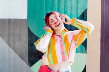 Emotional stylish young woman with pink hair in multicolor striped shirt wears wireless headphones, listening to music and singing on a graffiti wall background. enjoy the moment. Fashionable hipster. clipart