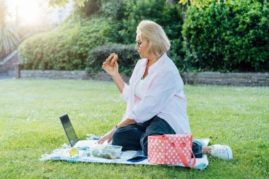 Orta yaşlı bir kadın yeşil çimlerde oturuyor, sağlıklı yemek yiyor ve dizüstü bilgisayarla çalışıyor. Uzaktan çalışmak, teneffüste piknik yapmak. Salata ve sandiviçle dengeli beslenme çantası. Sağlıklı beslenme alışkanlığı.